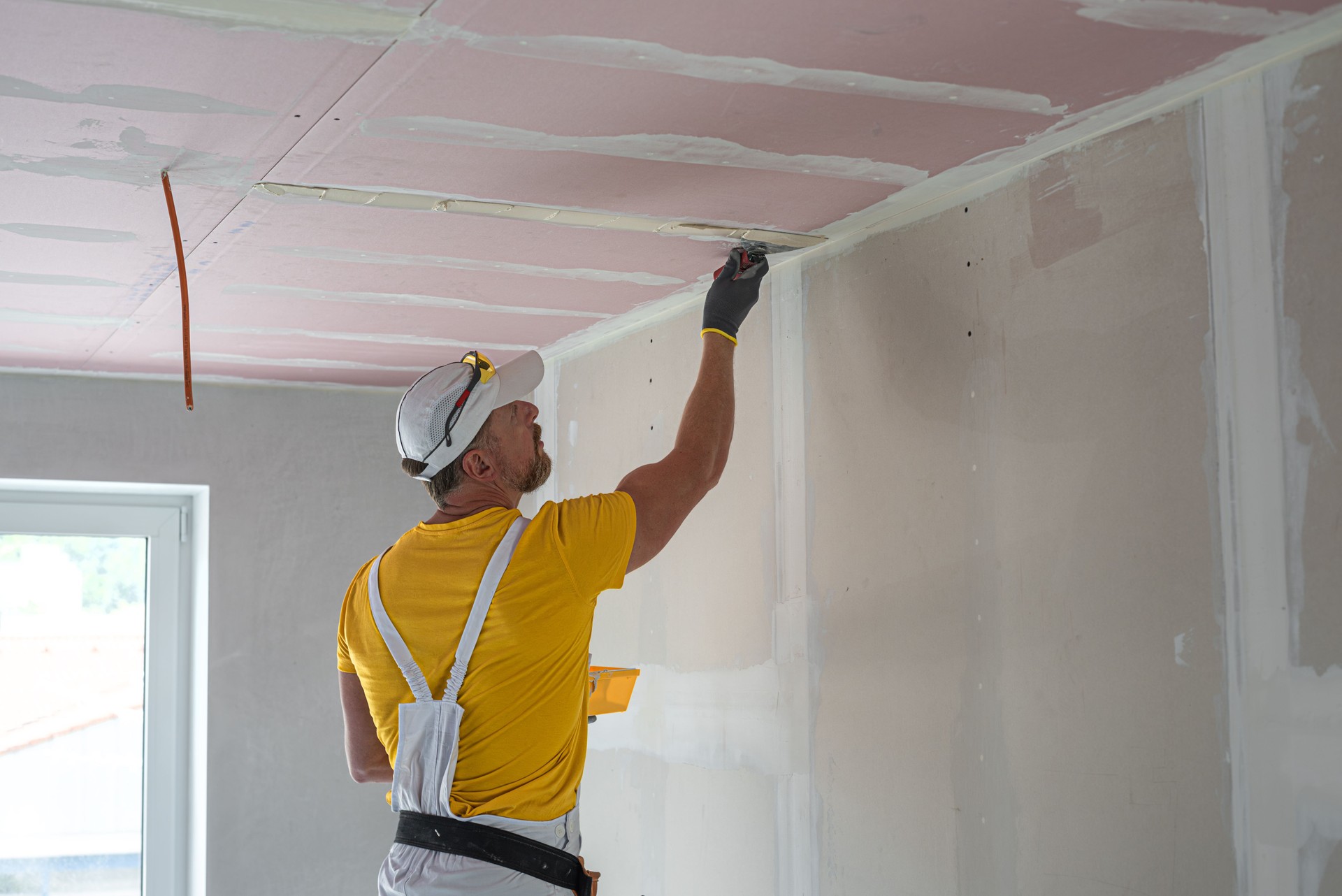 The worker make a plasterboard ceiling. He does taping plaster drywall ceiling joints. A red plasterboard improve the fire resistance of ceiling structures.